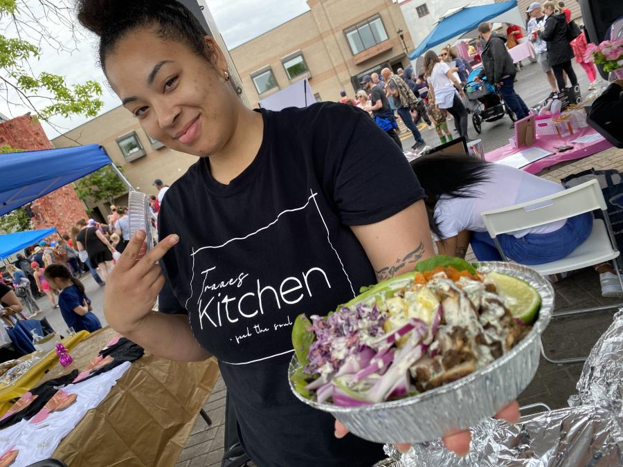 A black woman with a top bun wearing a black t shirt and holding a plate of colourful food.  she is a vendor at the apple blossom festival and is surrounded by people having fun in centre square, downtown.