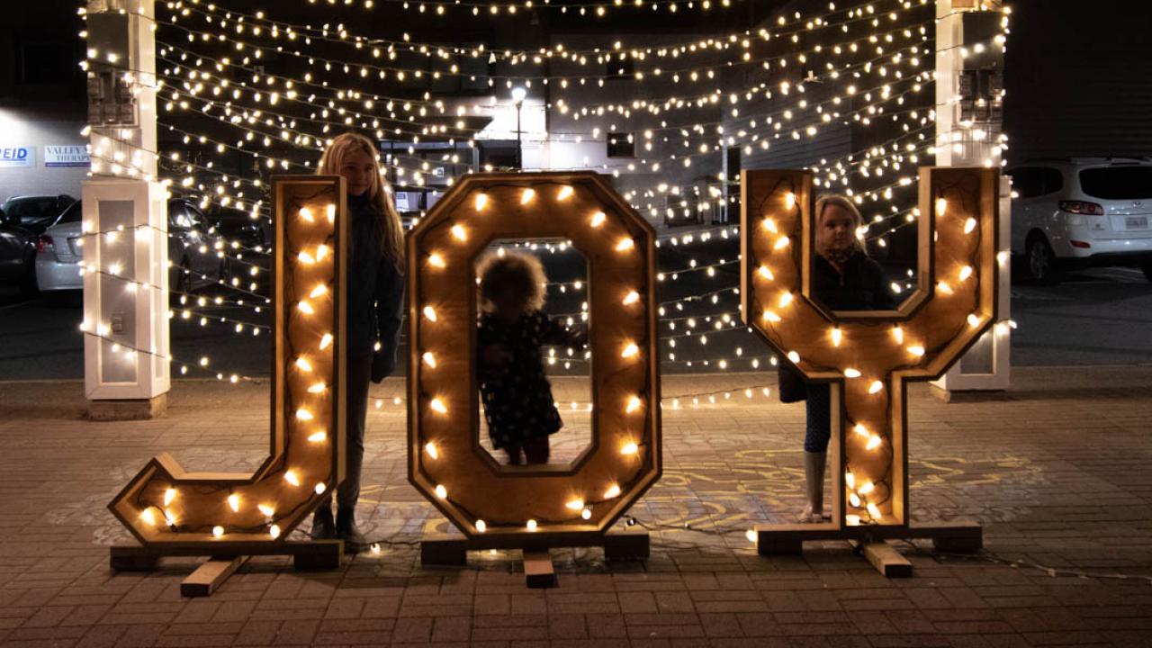 A family of three kids pose behind life-sized wooden letters that spell out the word joy. A backdrop of beautiful lights shines behind them. 