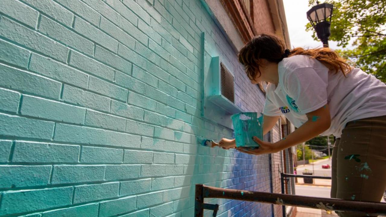 an artist with a long brown ponytail paints a blue and green base onto old brick 