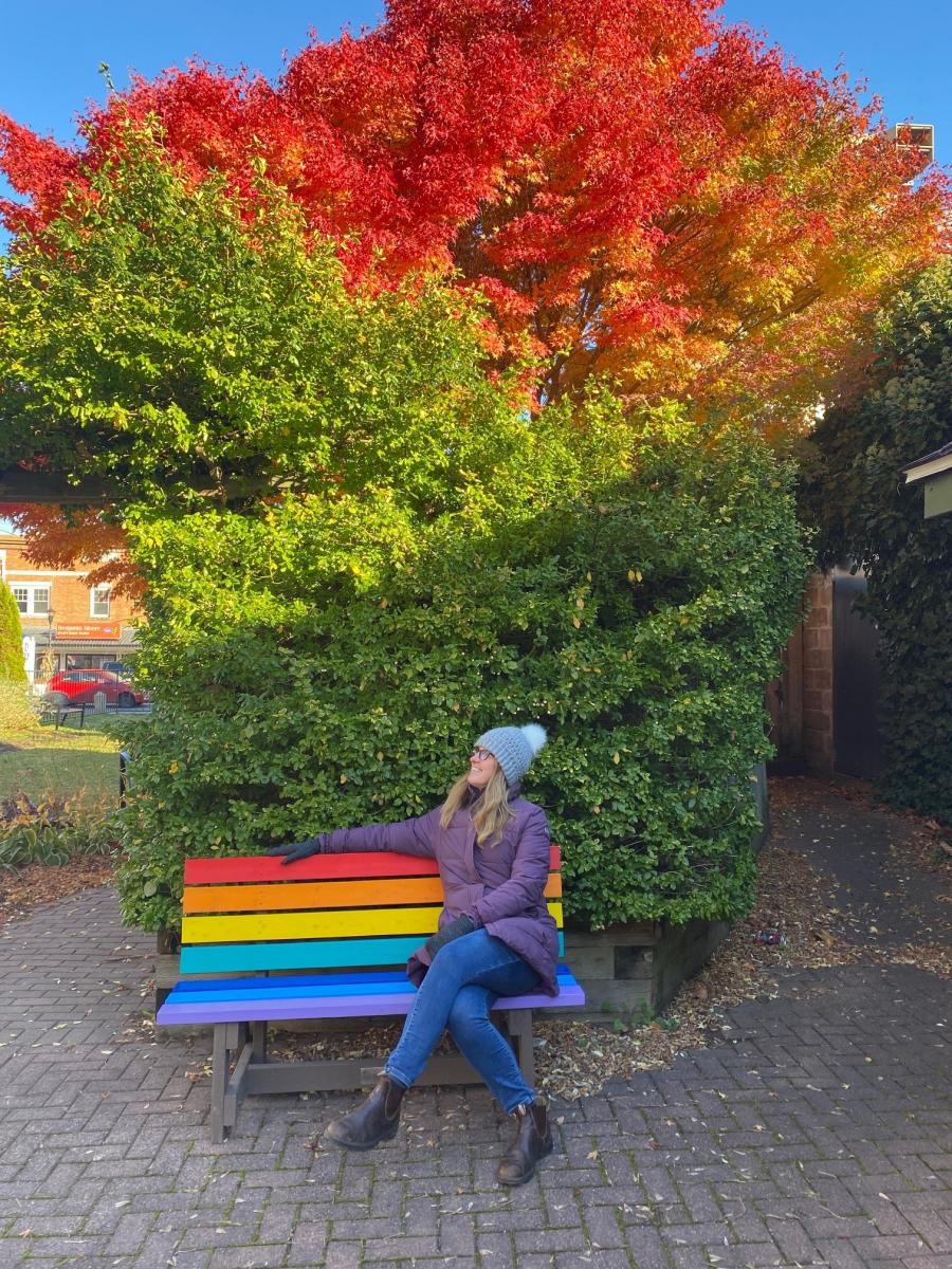 A woman sits on a rainbow coloured bench in front of a gorgeous japanese maple tree that is turning bright orange with the fall temperatures