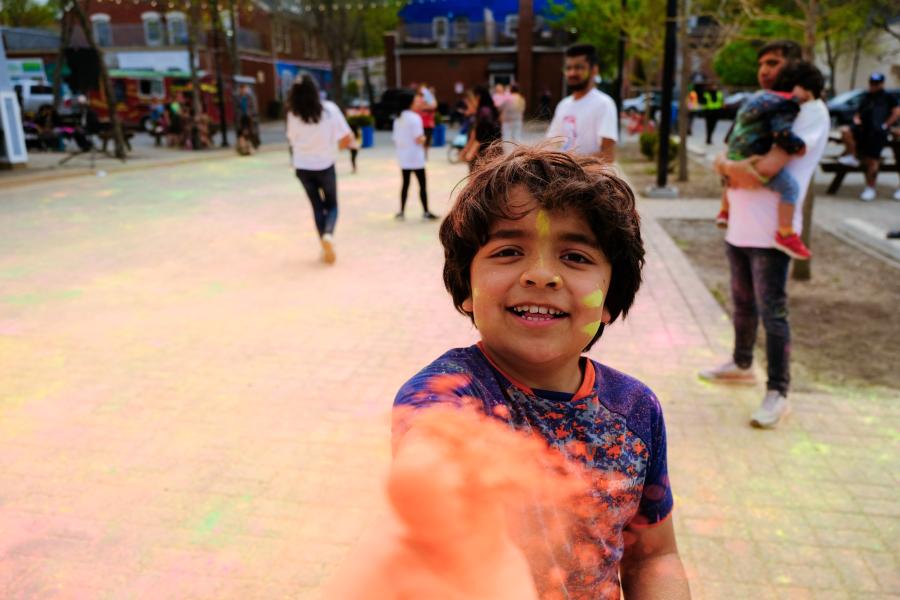 A kid covered in pigmented powder celebrtes Holi in downtown Kentville at Centre Square.  He is smiling as he holds out his hand toward the camera sharing the coloured powder and inviting you to join him at this year's Holi event. 