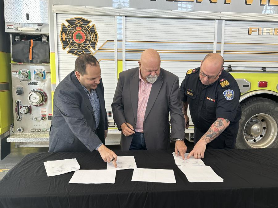 three men sign a contract at a black table with a firetruck in the background