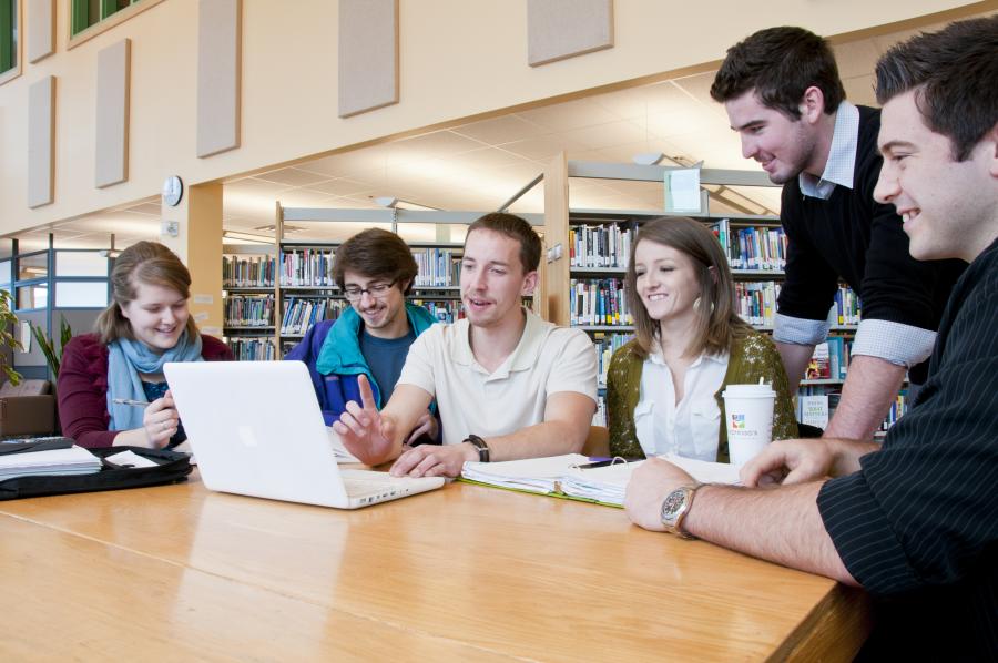 Six students working on a laptop around a table in the NSCC library.