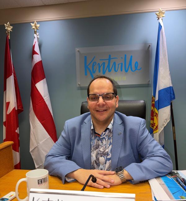 Mayor Andrew Zebian sitting at his seat in the Council Chambers, wearing glasses, a patterned shirt and a blue jacket. His official name tag is in the foreground and the Town logo is on the wall in the background in the middle of the Mi'kmaq Grand Council flag, the Canadian flag and the Nova Scotia provincial flag